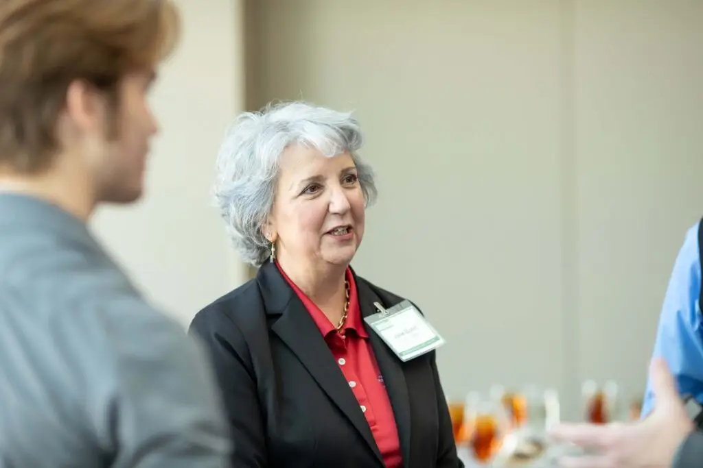 Older professional speaking with attendees during a networking event at a conference venue.